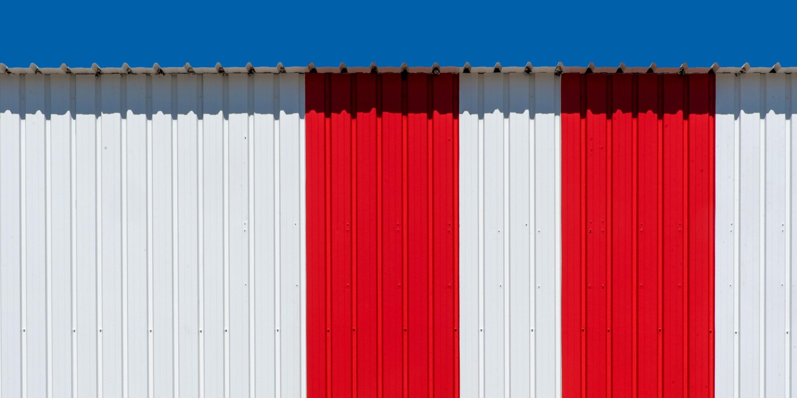 Minimalist photo of a red and white striped metal wall with a bright blue sky.