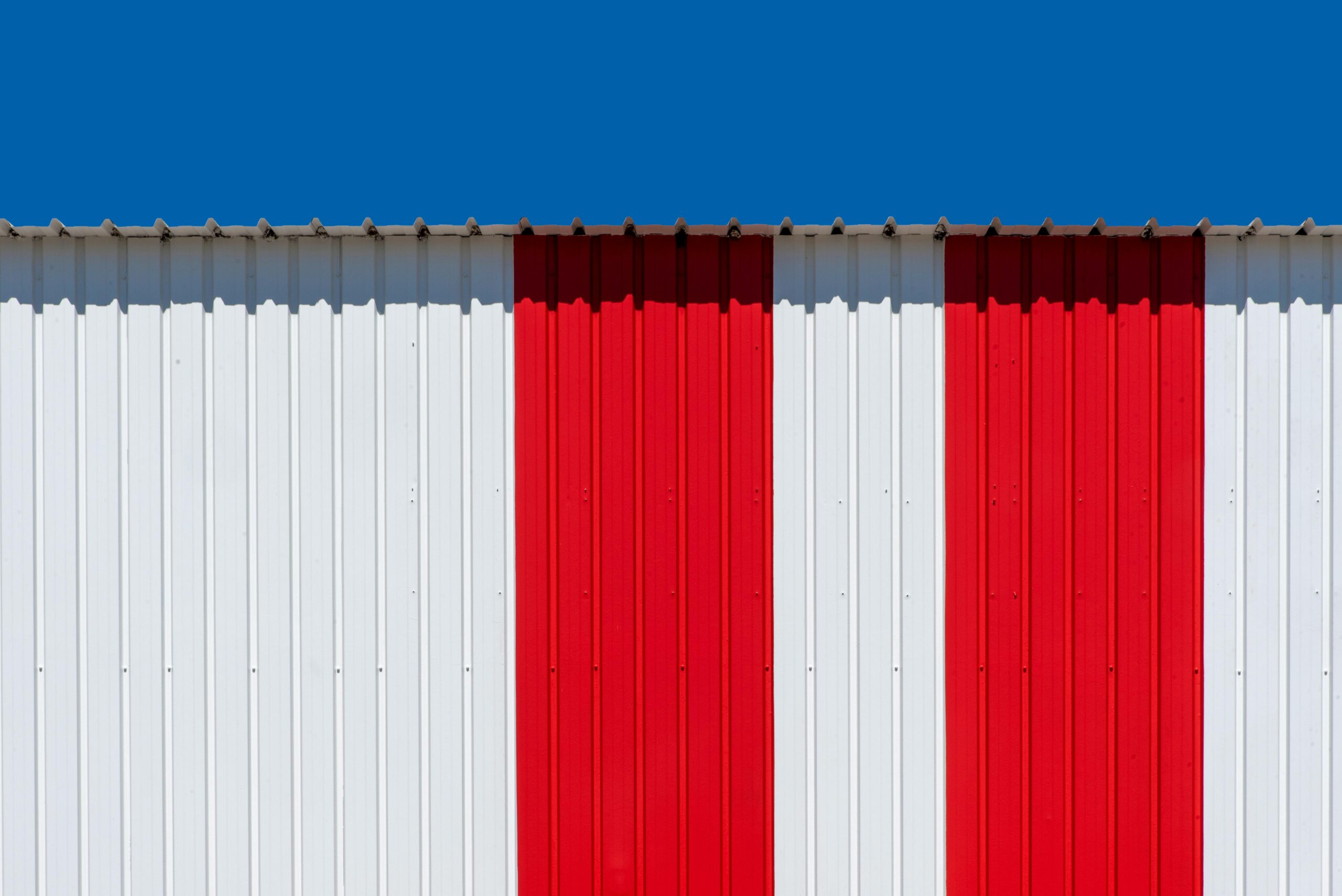 Minimalist photo of a red and white striped metal wall with a bright blue sky.
