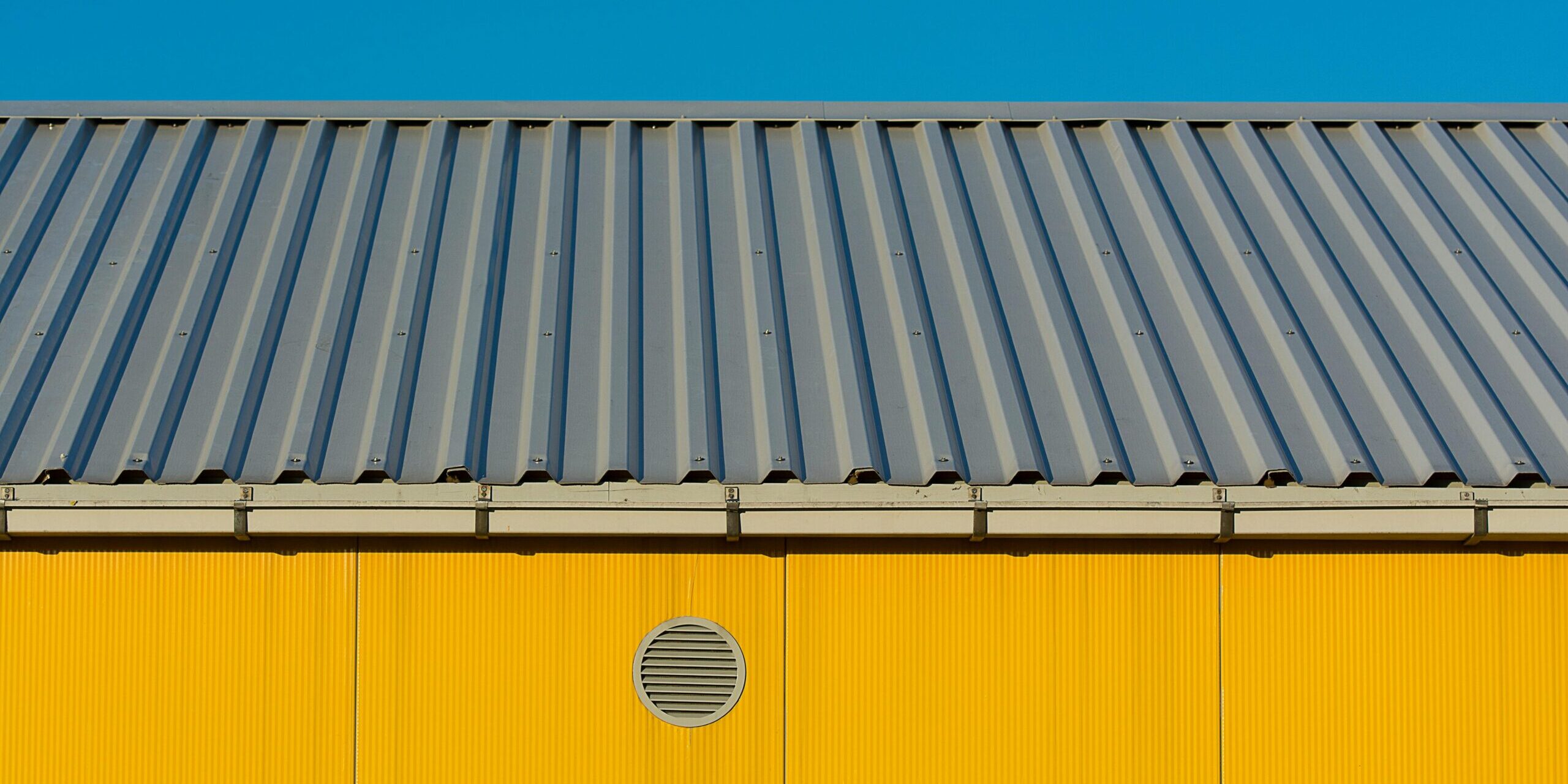 Modern industrial building’s yellow facade with me Modern industrial building's yellow facade with metal roof against clear blue sky.